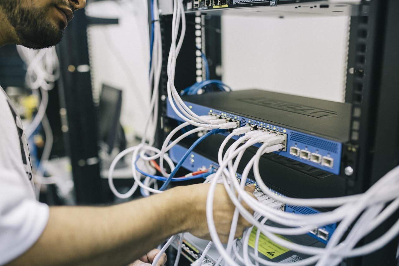 Un técnico con camiseta blanca, visto de espaldas y ligeramente de perfil, conecta cables Ethernet blancos en conmutadores de red azules montados en rack. Decenas de cables blancos cruzan la imagen, con algunos azules y uno naranja; abajo se ve una etiqueta de advertencia amarilla.