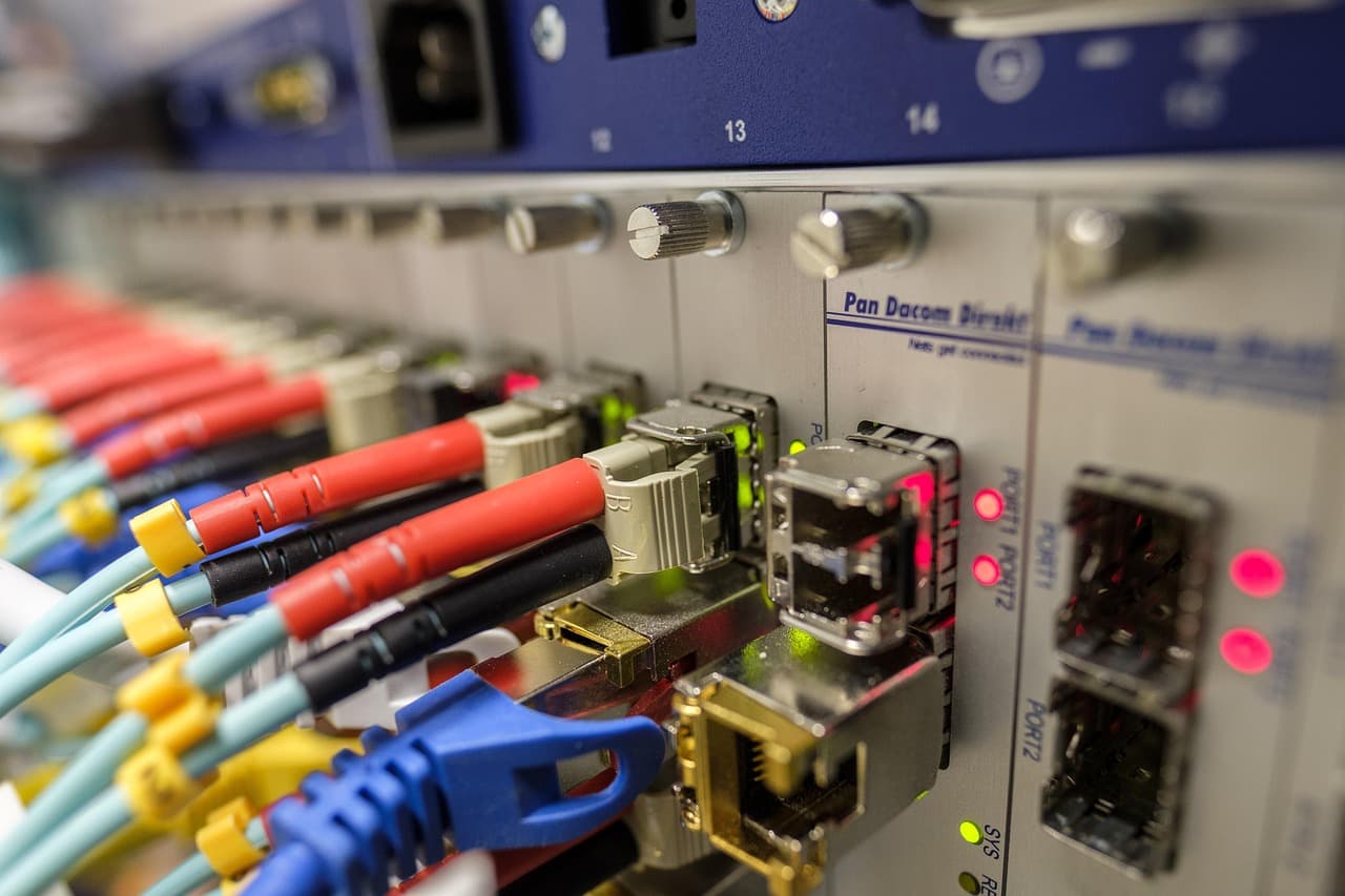 Panneau de brassage vu de près, jarretières rouges, turquoise, jaunes et bleues enfichées dans des modules étiquetés PORT1 et PORT2, voyants rouges et verts allumés