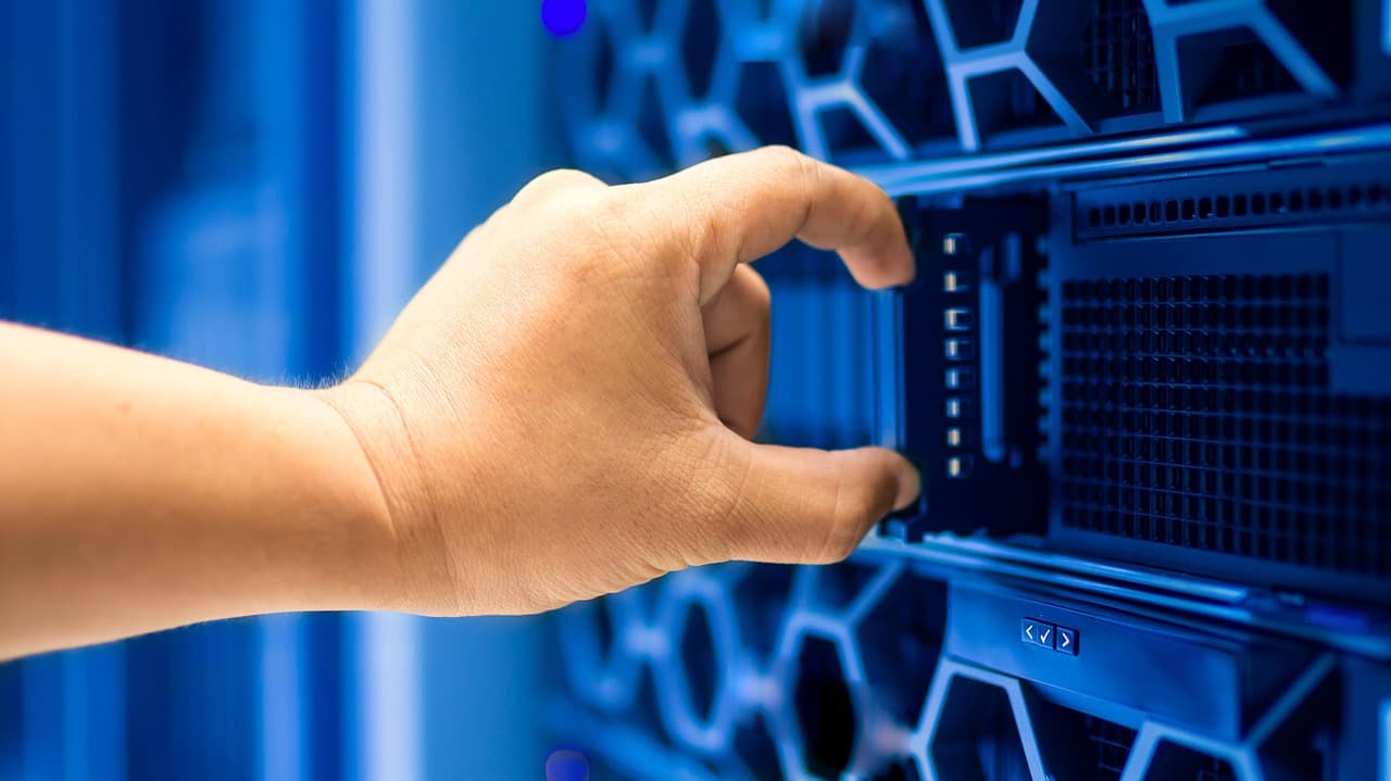 Une main extrait un tiroir de serveur d'une baie informatique éclairée en bleu, les grilles de ventilation en nid d'abeille remplissent l'arrière-plan