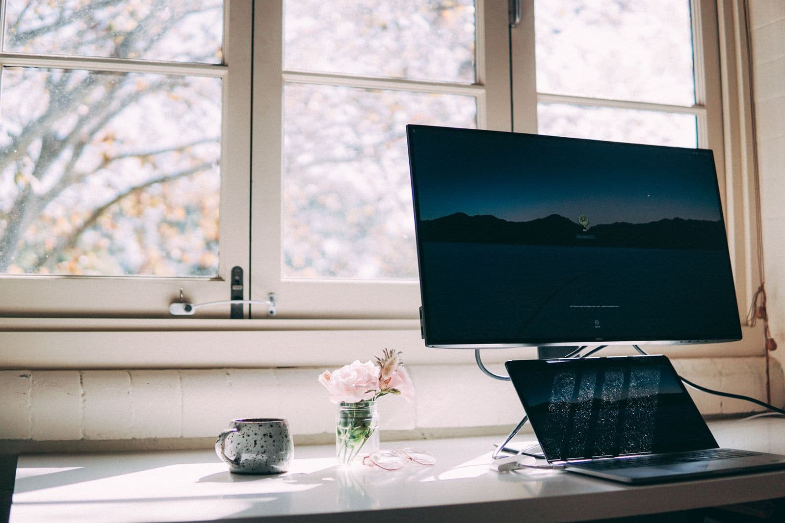 A desk in front of a bright multi-pane window: a large monitor showing a dark mountain-and-night-sky wallpaper with a sign-in prompt, an open laptop below it, a speckled grey mug, a small jar of pink flowers and a pair of glasses.