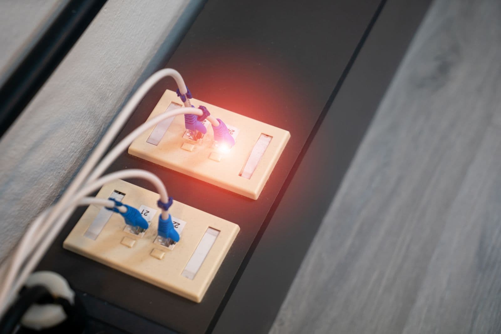 Two cream-coloured RJ45 wall sockets mounted on a dark panel, with four white Ethernet cables plugged in through blue and purple connector boots; a red LED glows on the upper socket, and the floor behind is pale grey.