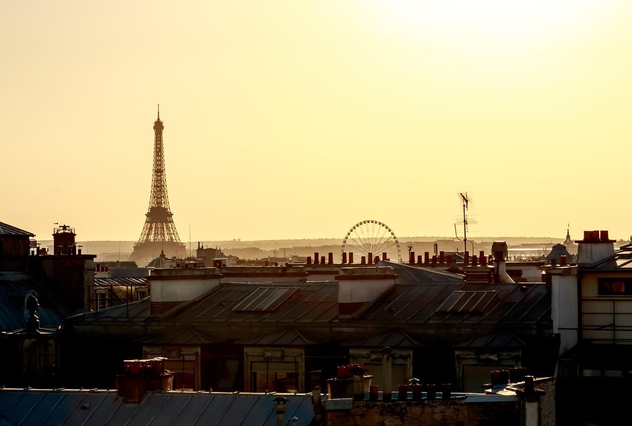 Toits de Paris au couchant, toitures de zinc et souches de cheminées au premier plan, tour Eiffel et grande roue en silhouette sur un ciel doré