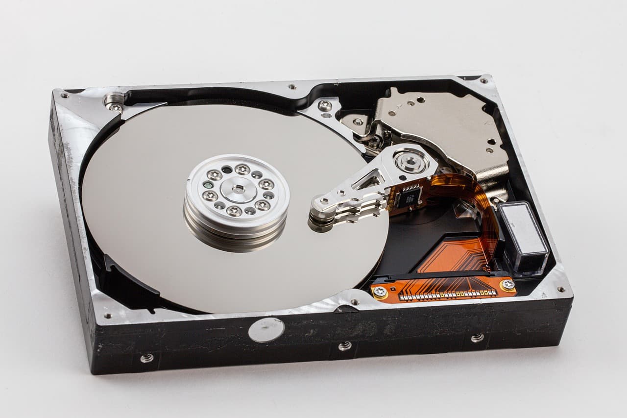 Un disque dur 3,5 pouces ouvert, capot retiré, plateau miroir et bras de lecture visibles, nappe orange sur le circuit, posé sur fond blanc.