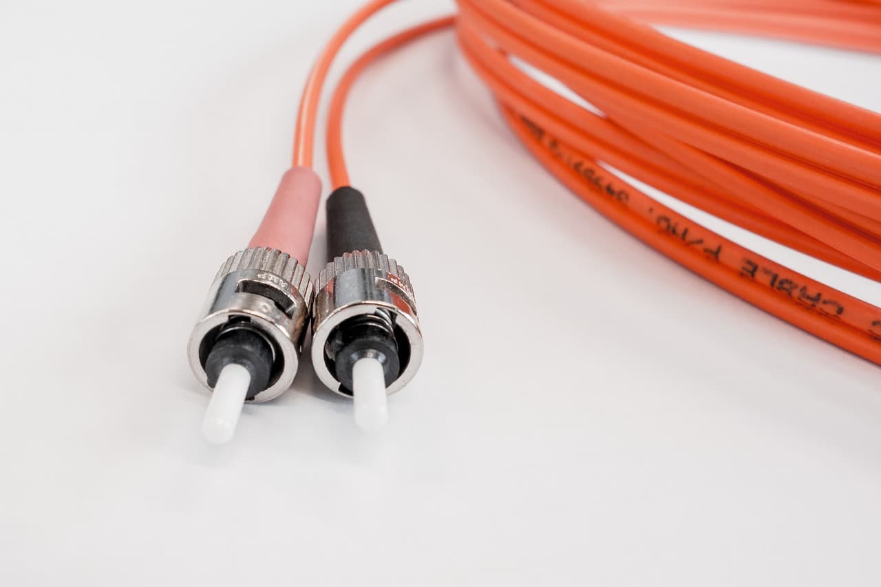 Dois cabos de fibra ótica laranja com conectores metálicos ST, o cabo enrolado etiquetado ao fundo