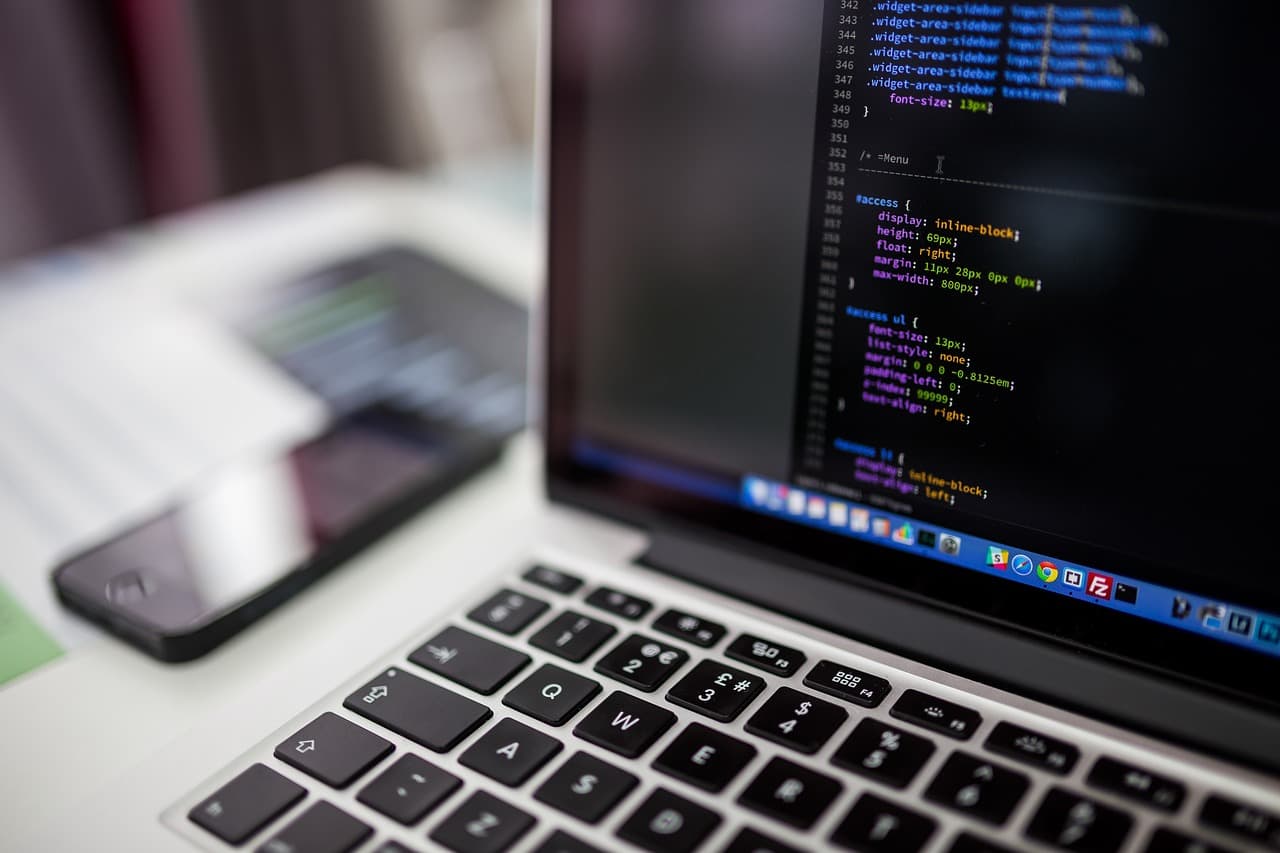 Un laptop su una scrivania che mostra righe di codice sorgente in un editor scuro, con uno smartphone appoggiato accanto alla tastiera